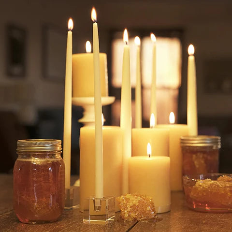 Beeswax taper and pillar candles lighting up a dim room. There is also jars of honey and honeycomb in the photo.