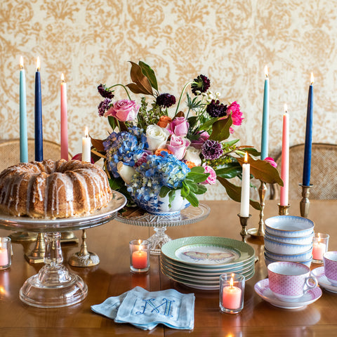 Dining table set with cake, flowers, taper and pillar candles, and tea sets against a patterned wall.